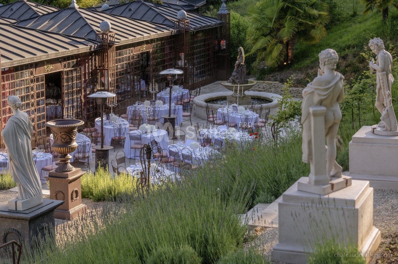Terrasse élégante avec tables dressées, statues classiques et fontaine entourée de verdure.