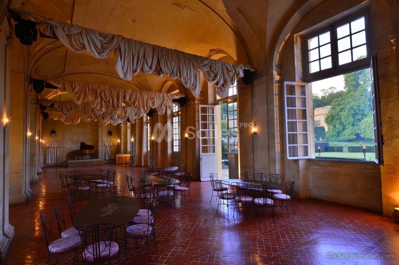 Salle spacieuse avec tables en fer forgé, rideaux suspendus, grandes fenêtres ouvertes sur un jardin verdoyant.