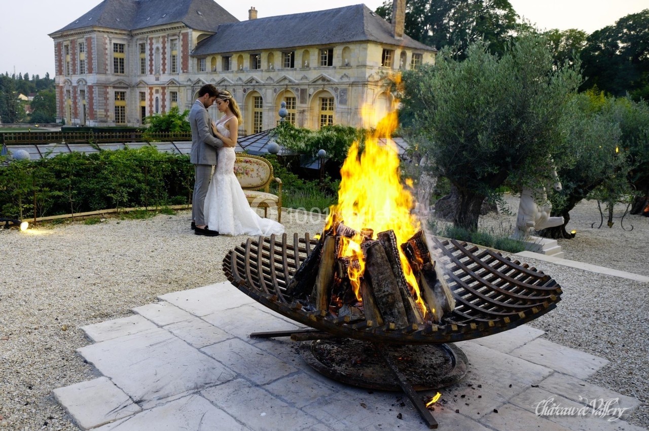 Un couple en tenue de mariage s'embrasse près d'un brasero allumé, devant un château ancien.