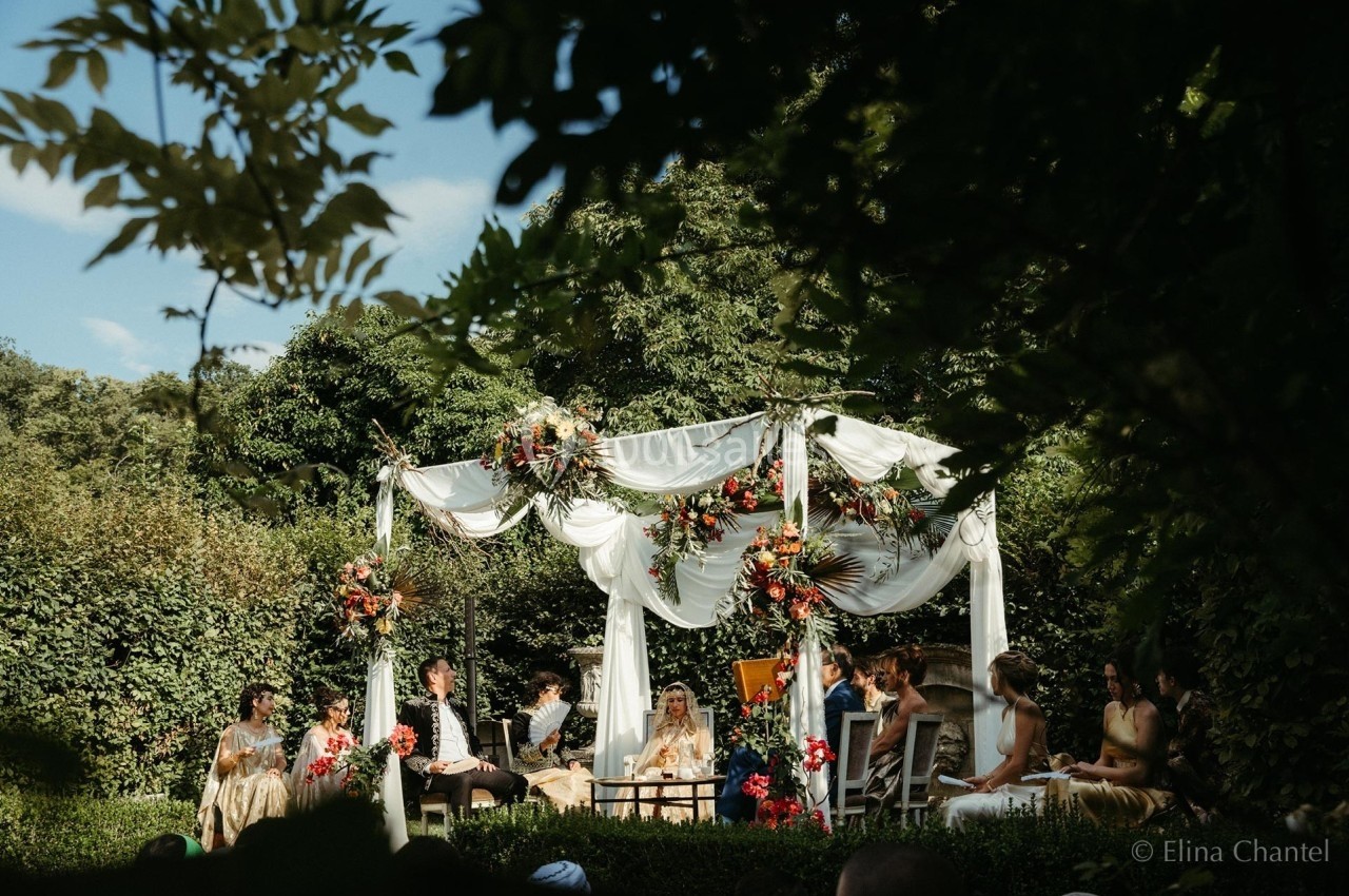 Cérémonie de mariage en extérieur sous une pergola décorée de fleurs, entourée de verdure et d'invités assis.