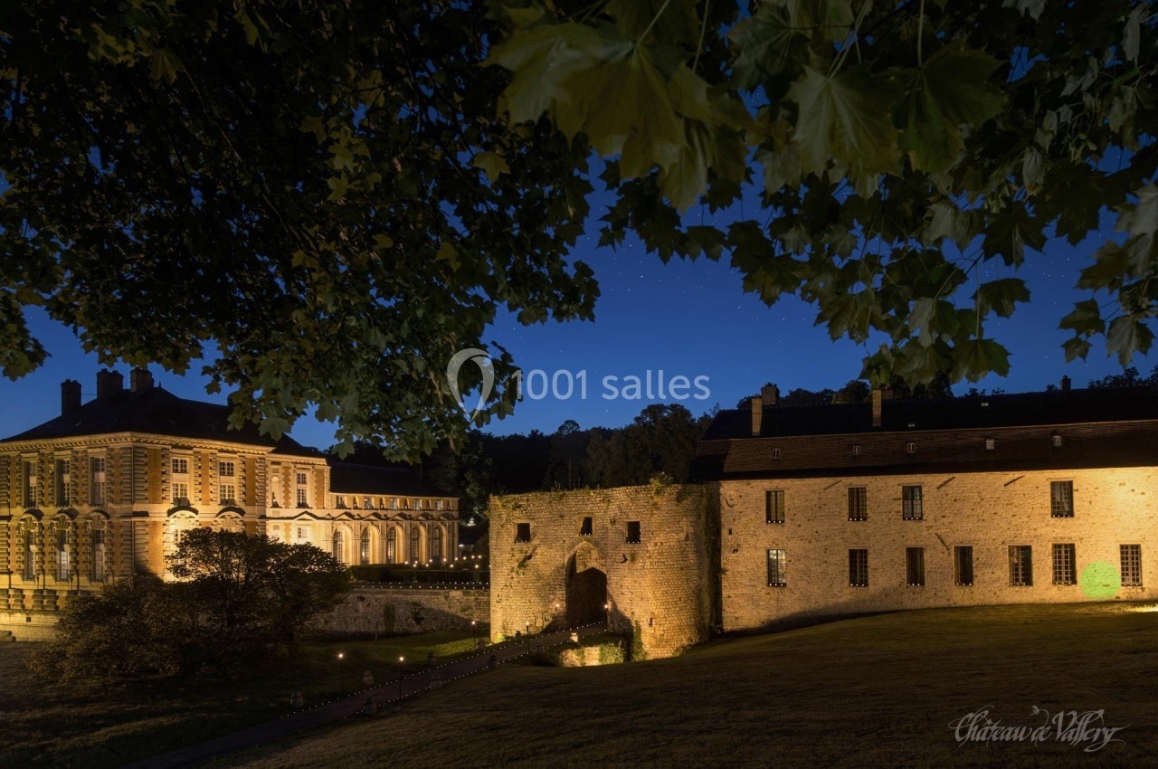 Vue nocturne d'un château éclairé avec une cour, des bâtiments historiques et des arbres au premier plan.