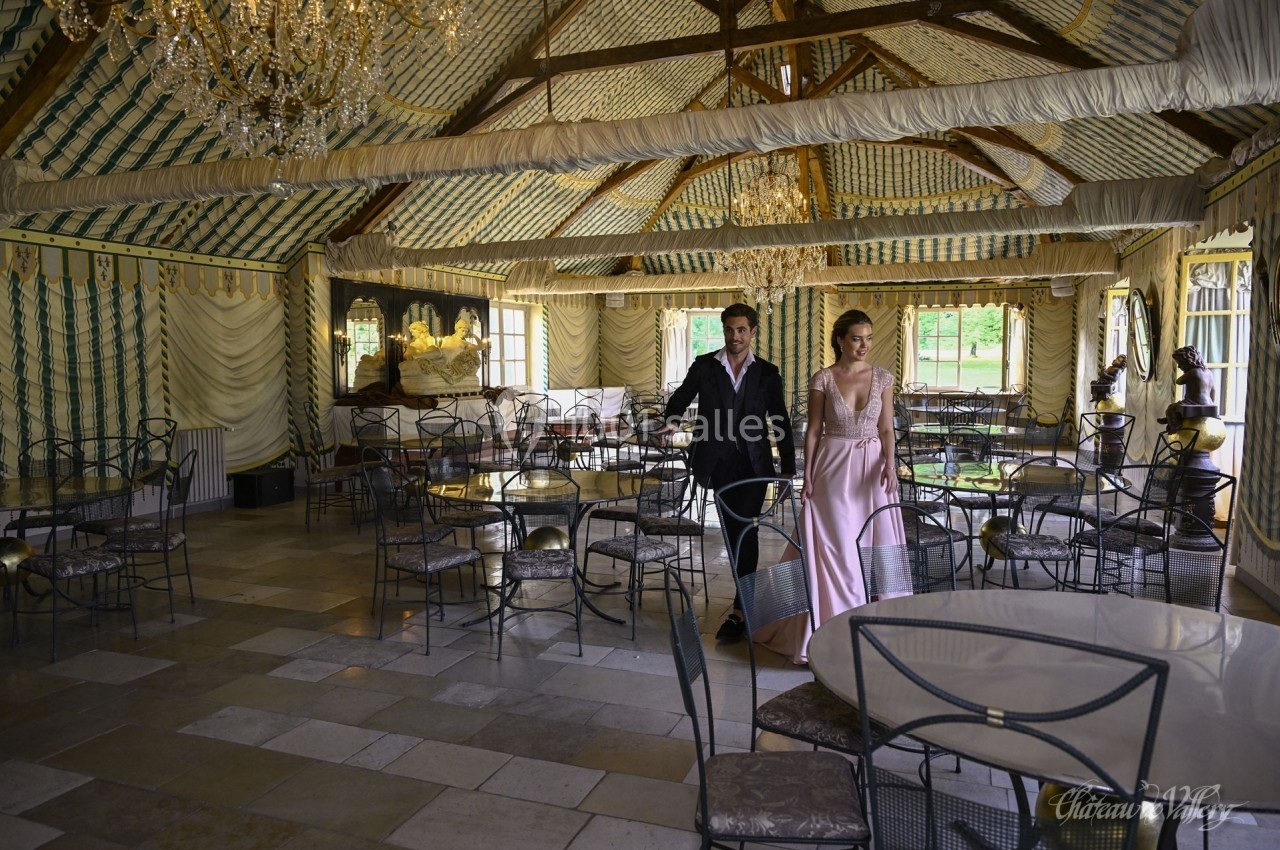 Un couple élégant marche dans une salle de réception décorée avec des lustres et des tables en fer forgé.