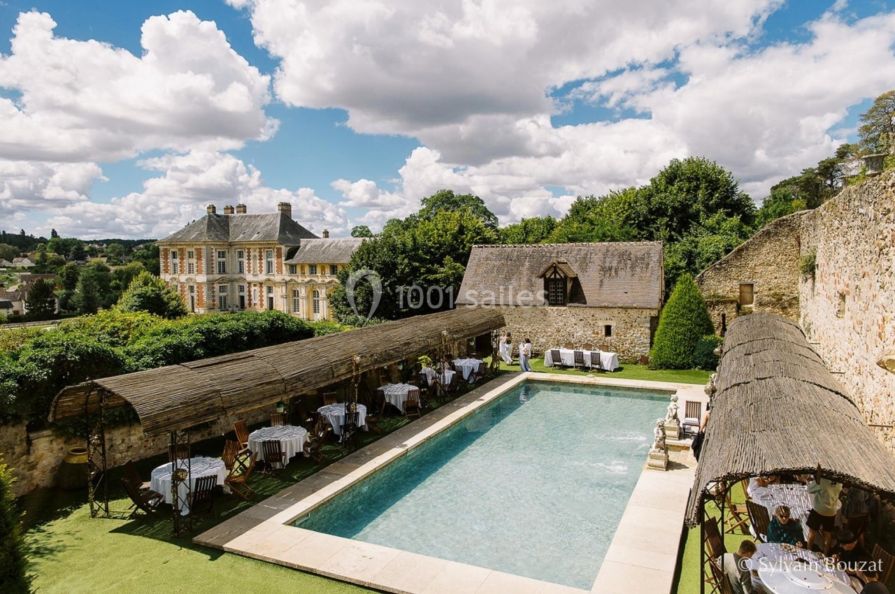 Vue d'une piscine extérieure entourée de pergolas, tables dressées et bâtiments anciens sous un ciel partiellement nuageux.
