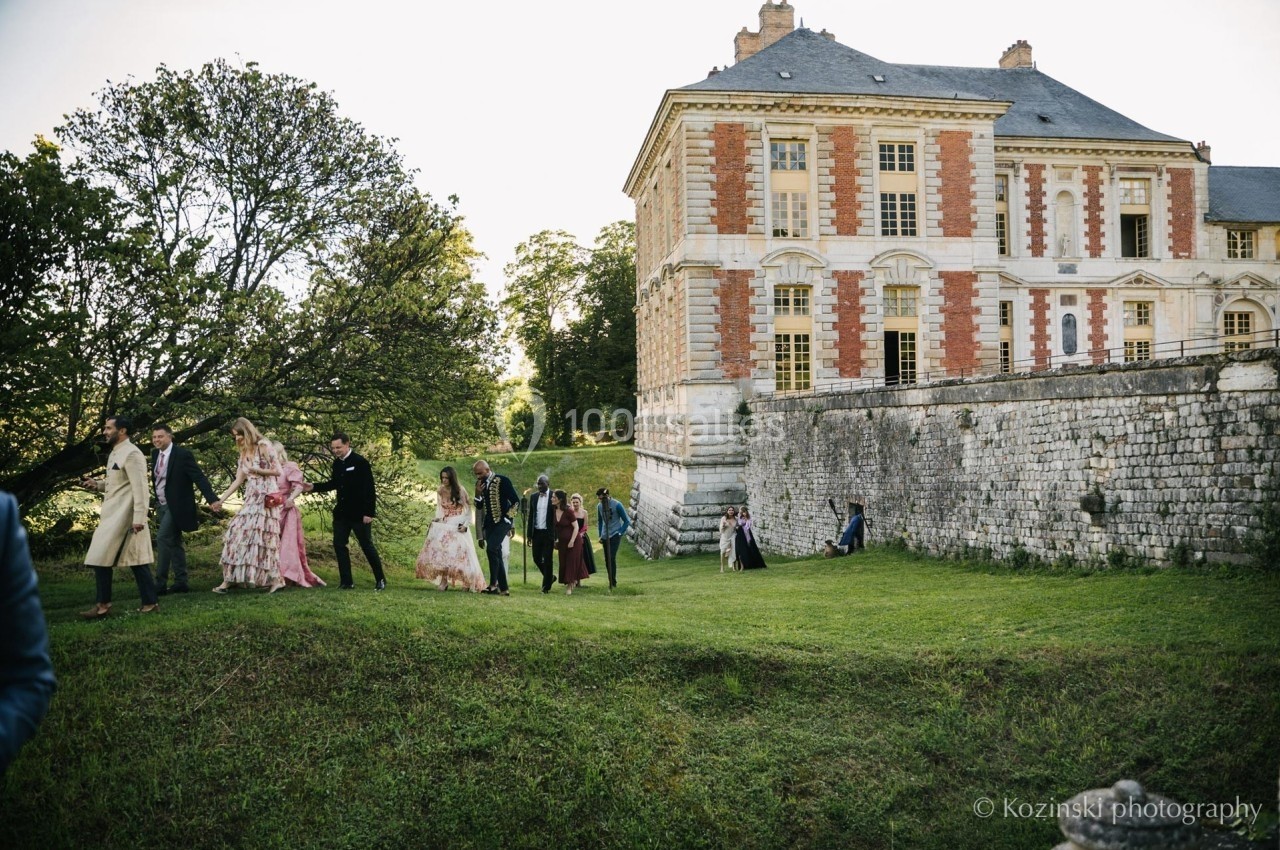 Un groupe de personnes élégamment vêtues marche sur une pelouse près d'un château en pierre et briques rouges.