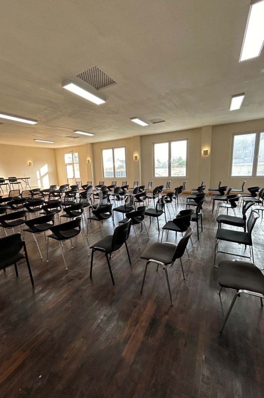 Salle lumineuse avec parquet sombre, rangées de chaises noires vides et grandes fenêtres laissant entrer la lumière…
