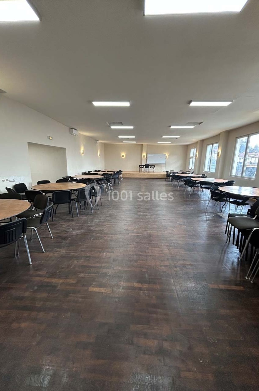 Salle spacieuse avec parquet sombre, tables rondes et chaises disposées, éclairée par des fenêtres et des plafonniers.