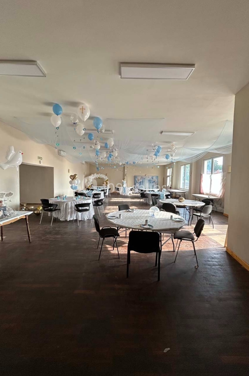Salle décorée pour une fête avec ballons bleus et blancs, tables dressées et lumière naturelle entrant par les fenêtres.