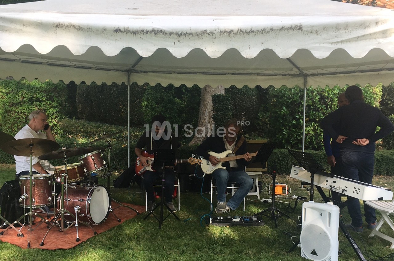 Musiciens jouant sous une tente blanche dans un jardin, avec batterie, guitares et clavier.