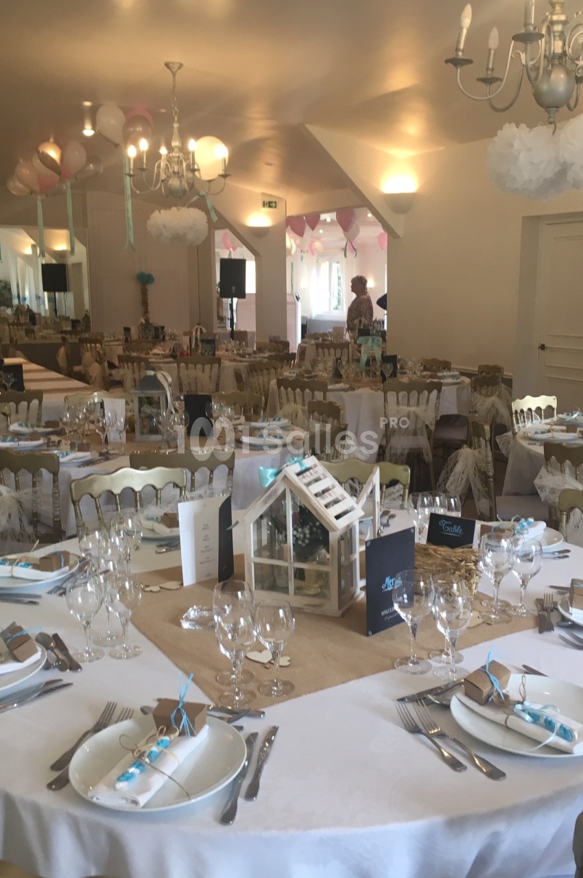 Salle décorée pour un événement avec tables dressées, nappes blanches, couverts élégants et décorations suspendues.