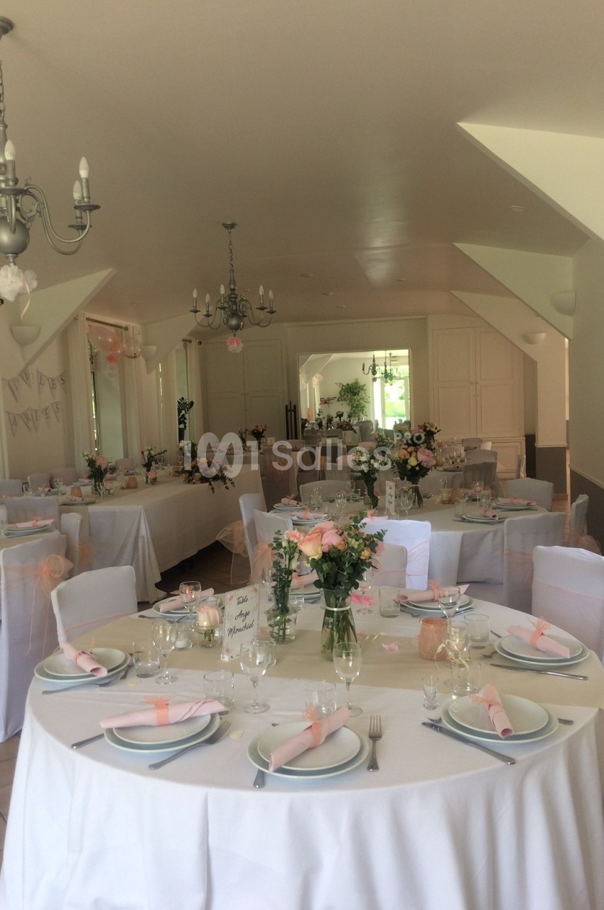 Salle de réception décorée avec des tables dressées, nappes blanches, fleurs pastel et chaises ornées de rubans roses.