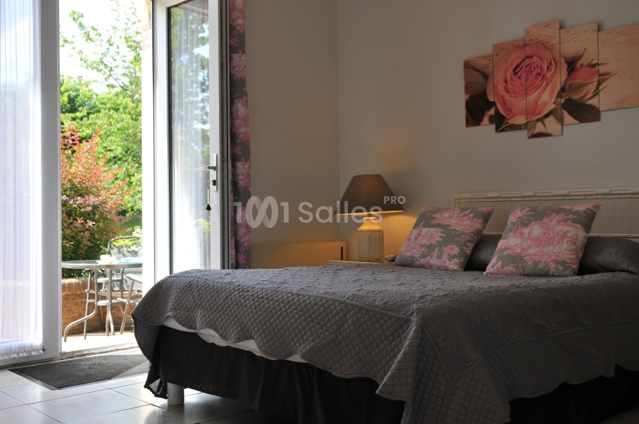 Chambre lumineuse avec lit double, décoration florale, lampe de chevet et accès à une terrasse avec table extérieure.