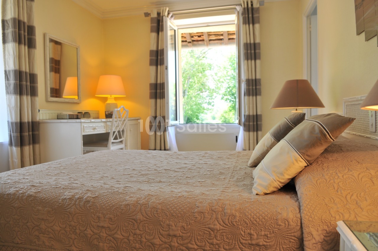 Chambre lumineuse avec un lit double, des lampes de chevet, un bureau blanc et une fenêtre ouverte sur un jardin.