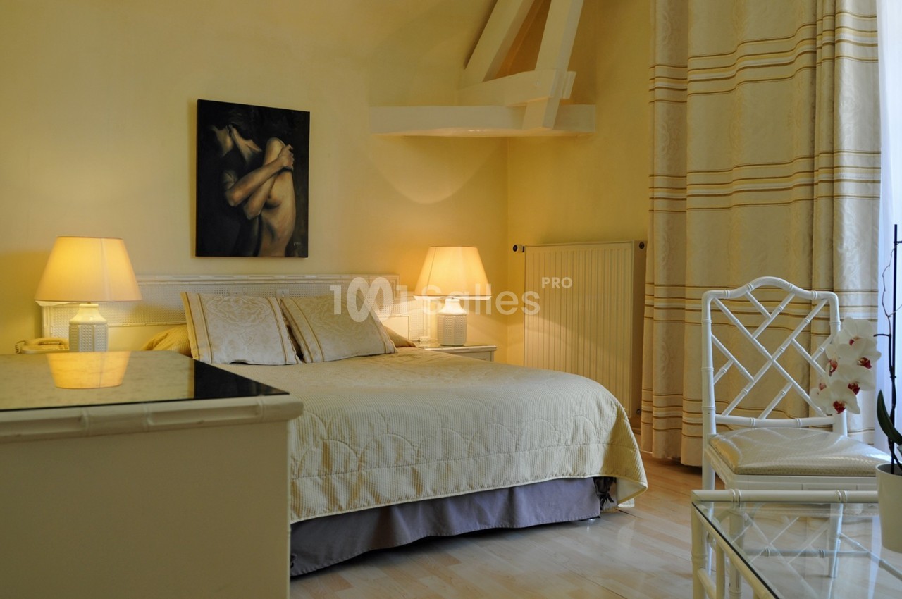 Chambre lumineuse avec lit double, murs jaunes, lampes de chevet, tableau, orchidée et grande fenêtre avec rideaux.