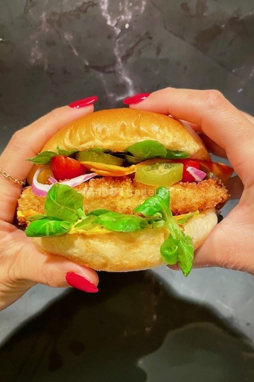 Deux mains tiennent un burger garni de salade, cornichons, oignons rouges et un ingrédient pané.