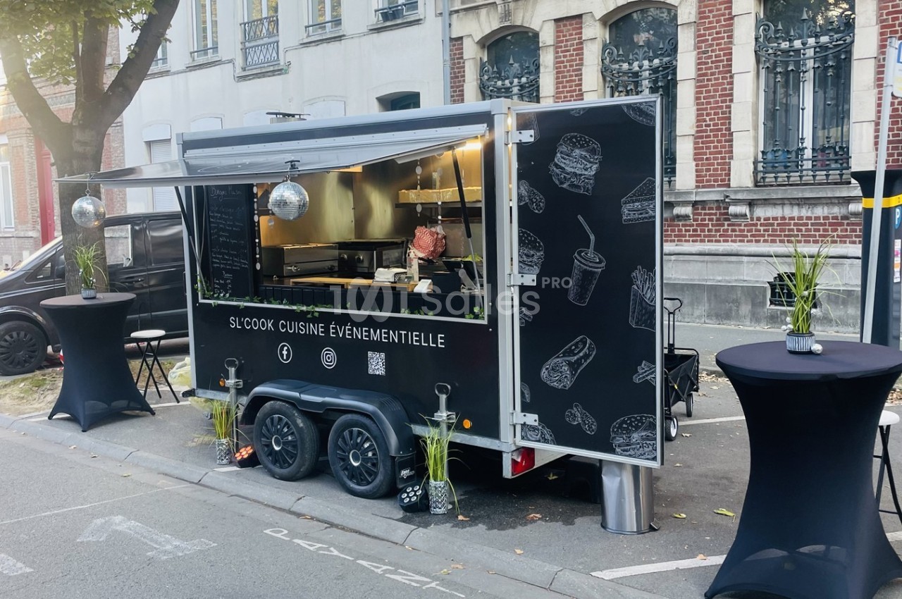 Food truck noir stationné dans une rue, avec des tables hautes et des illustrations de plats sur sa façade.
