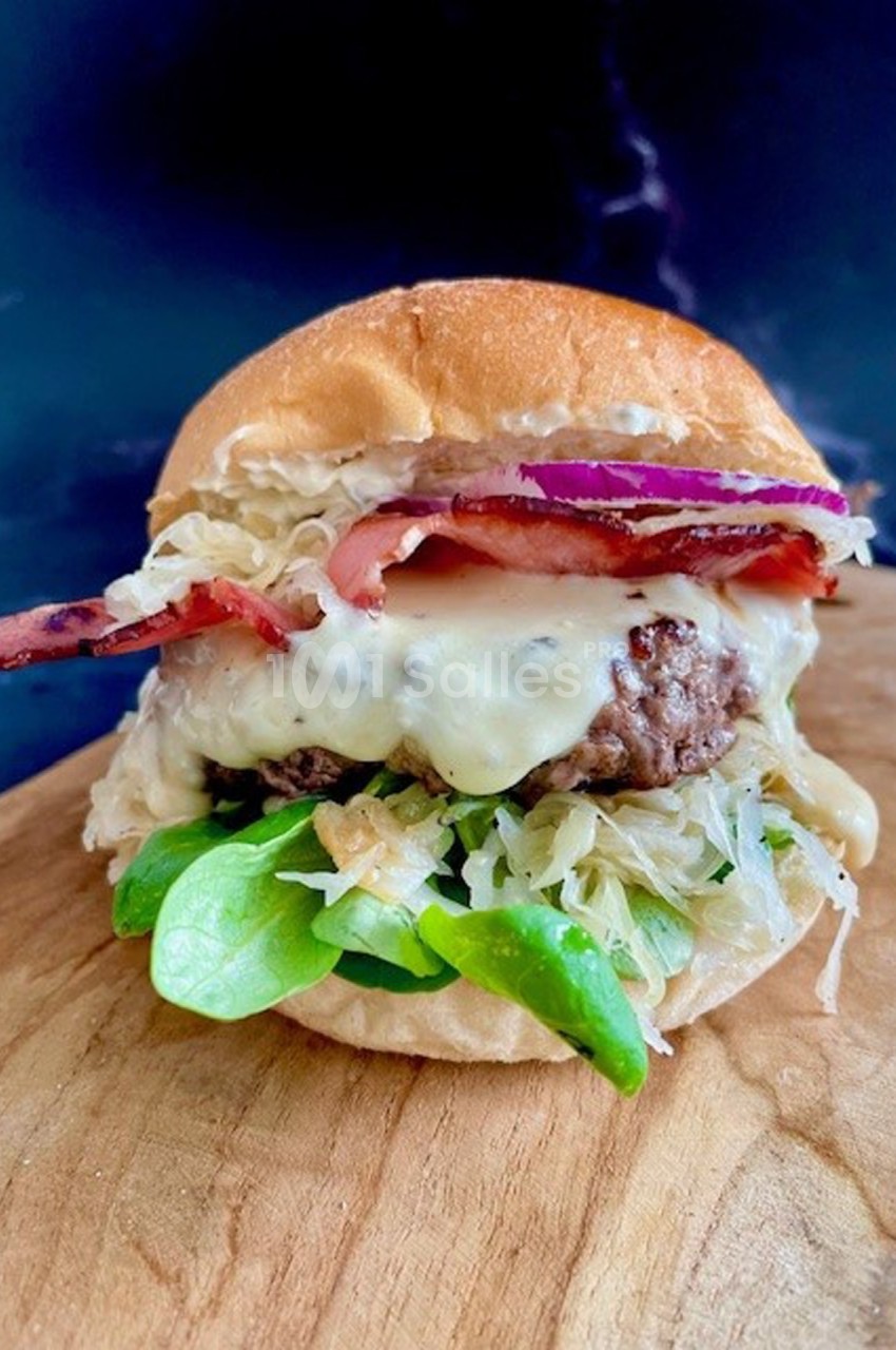 Burger garni de fromage fondu, bacon, oignons rouges, salade et choucroute, posé sur une planche en bois.