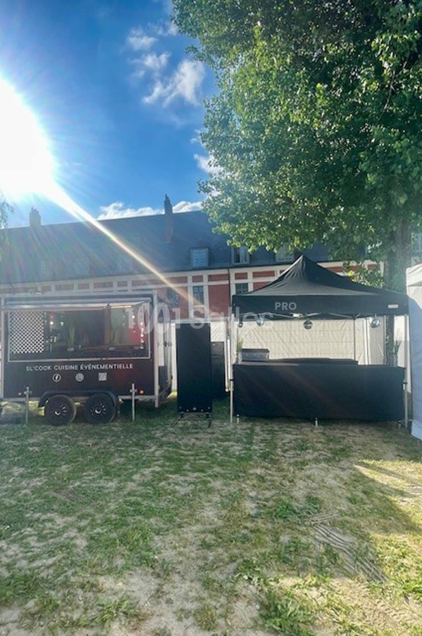 Camion de cuisine événementielle et stand noir installés sur une pelouse, avec des arbres et un bâtiment en arrière-plan.