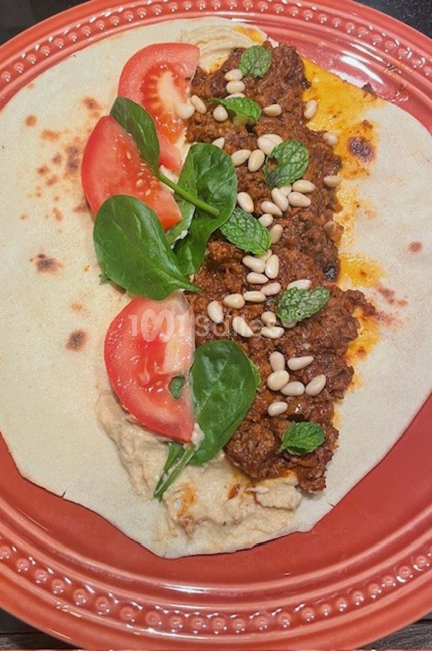 Wrap ouvert garni de viande épicée, tomates, feuilles d'épinard, pignons de pin, menthe et houmous sur une assiette rouge.