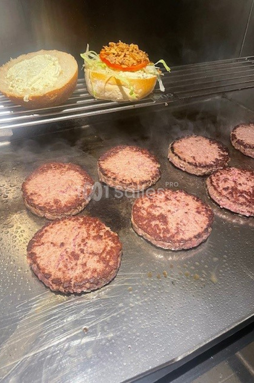 Burgers en cuisson sur une plaque chauffante avec un pain garni de légumes et sauce en arrière-plan.