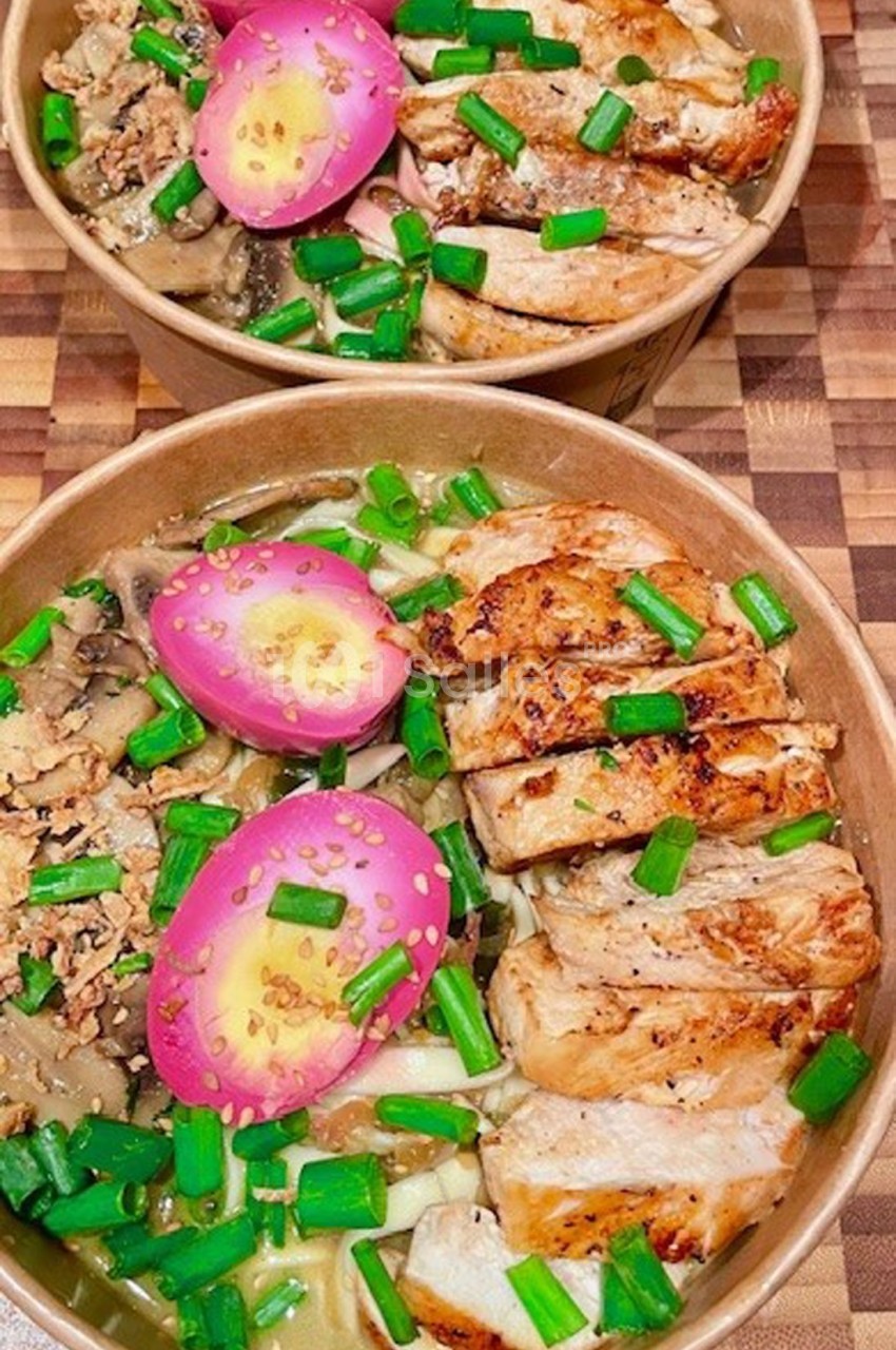 Deux bols de ramen garnis de poulet grillé, œufs marinés roses, champignons et ciboulette sur une table en bois.