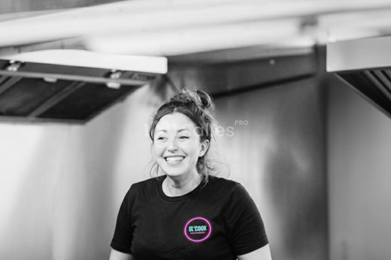 Femme souriante portant un t-shirt noir et des gants, travaillant dans une cuisine professionnelle.
