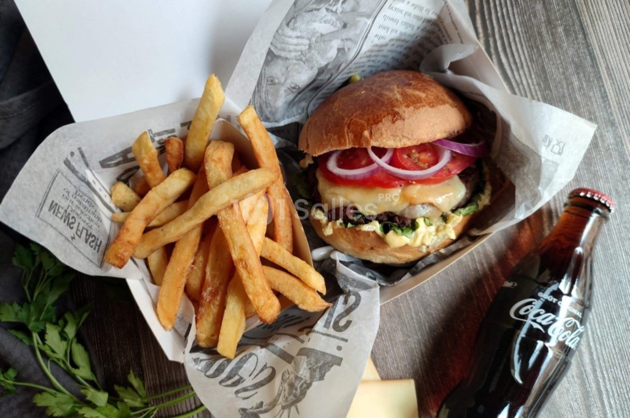 Burger garni avec frites dans un emballage en papier, accompagné d'une bouteille de Coca-Cola sur une table en bois.
