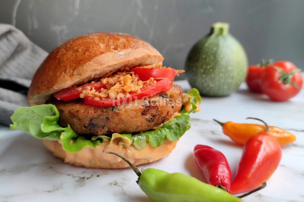 Burger végétarien garni de salade, tomate et oignons frits, avec des légumes frais en arrière-plan sur un plan de travail.