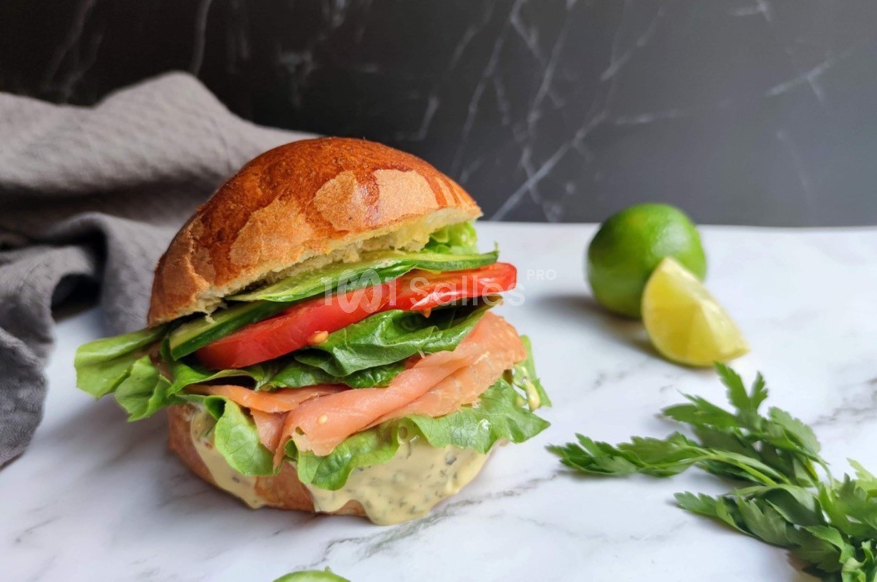 Sandwich garni de saumon fumé, laitue, tomate et sauce, posé sur une table en marbre avec citron vert et herbes.