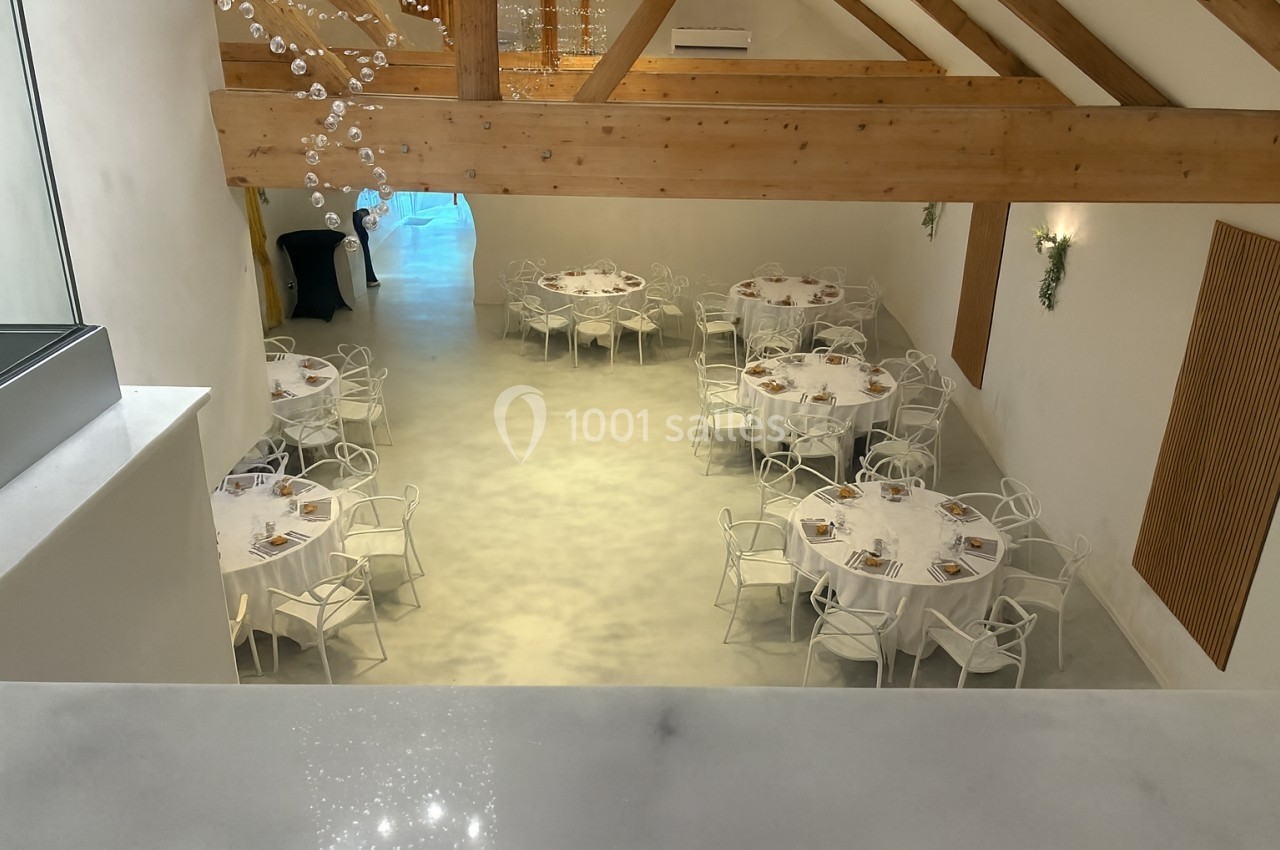 Salle lumineuse avec tables rondes dressées pour un événement, poutres apparentes et décorations minimalistes.