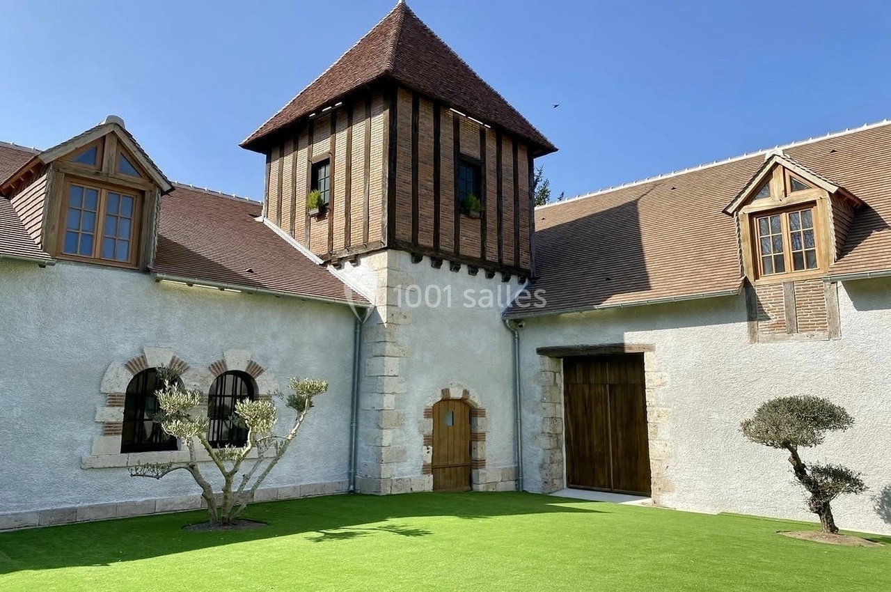 Bâtiment ancien avec tour centrale en briques, toit en tuiles et façade blanche, entouré d'une cour en gazon.