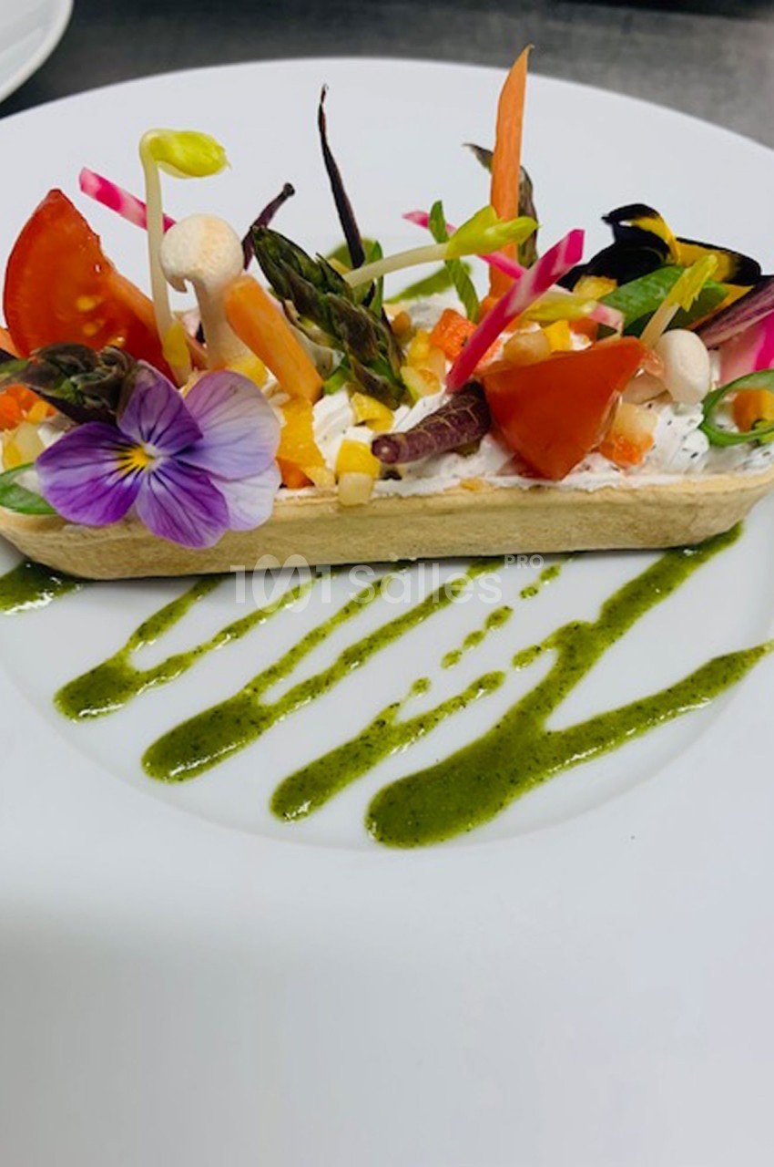 Tartelette garnie de légumes colorés, fleurs comestibles et sauce verte disposée en traits sur une assiette blanche.