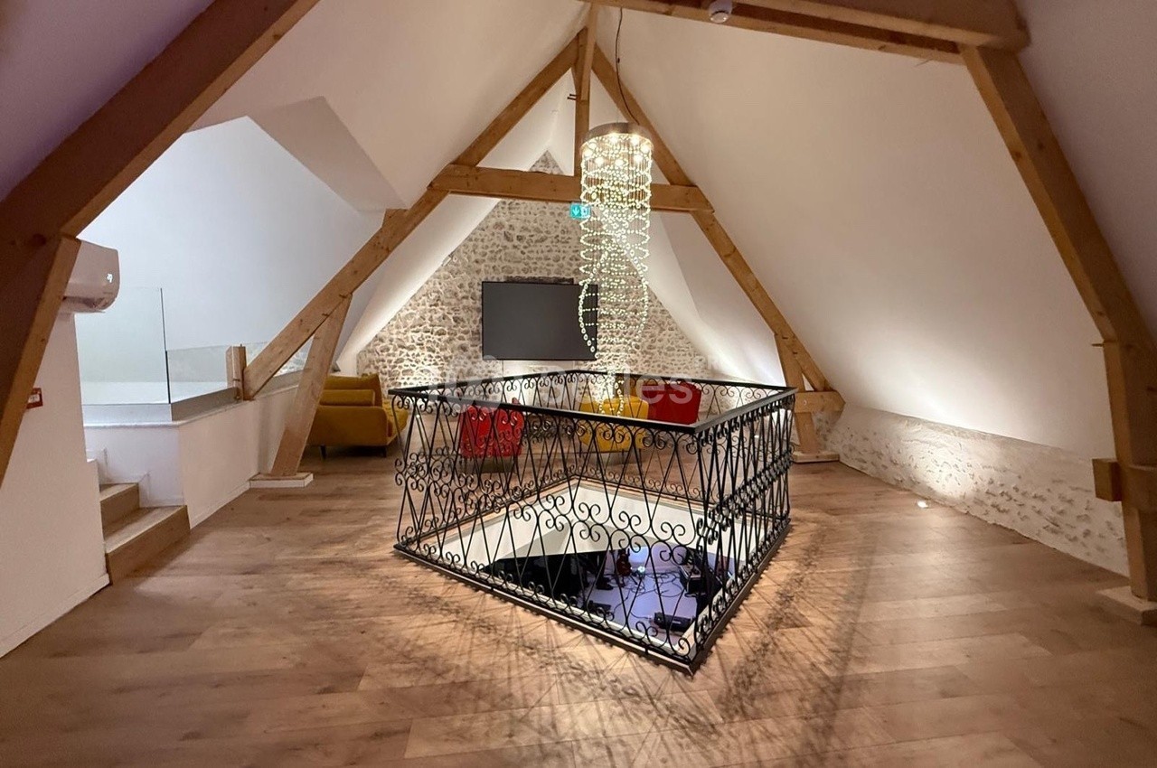 Grenier aménagé avec parquet, mur en pierre, garde-corps en fer forgé et lustre moderne, espace lumineux et chaleureux.