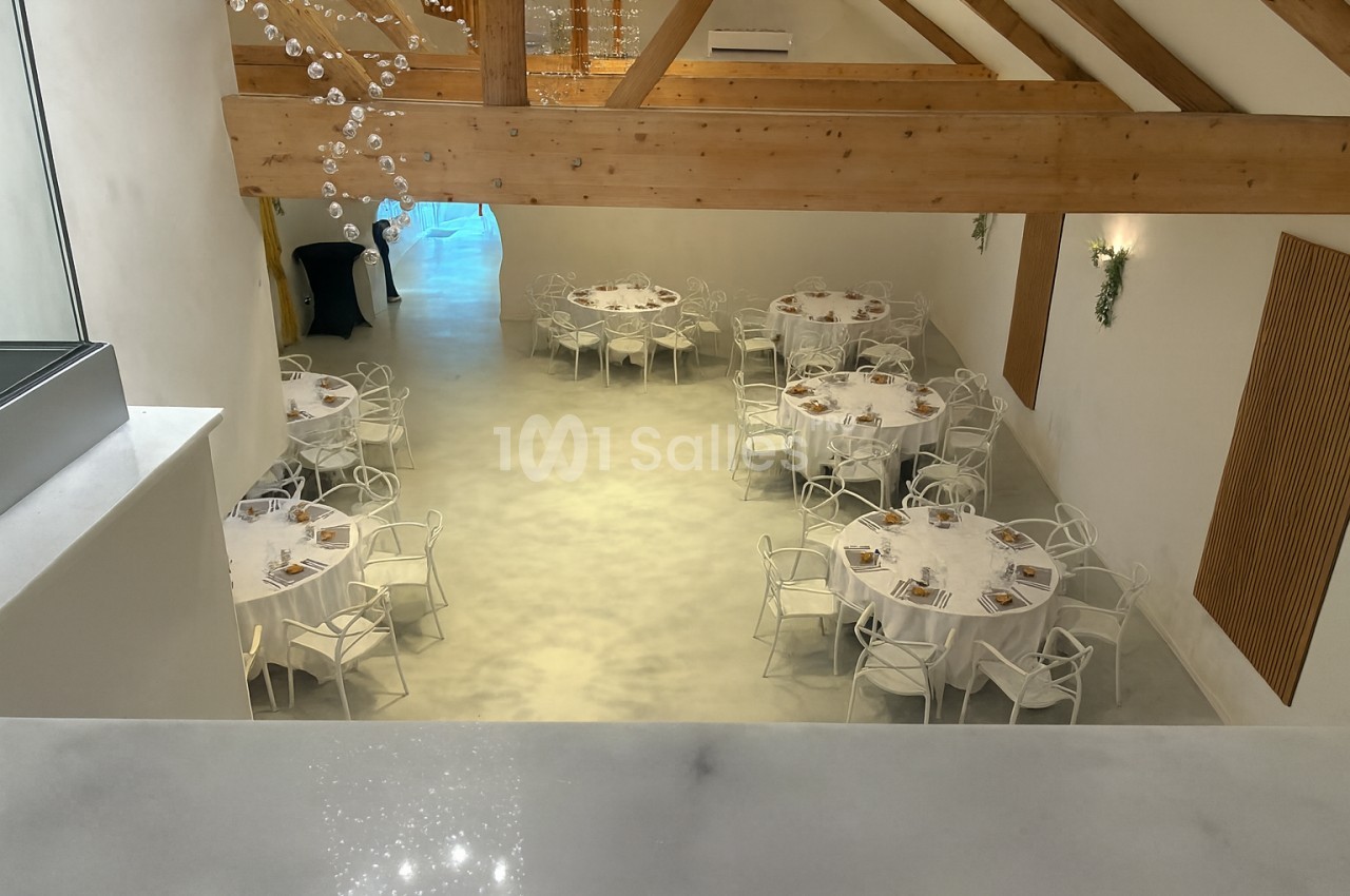 Salle lumineuse avec tables rondes dressées, chaises blanches et poutres apparentes en bois.