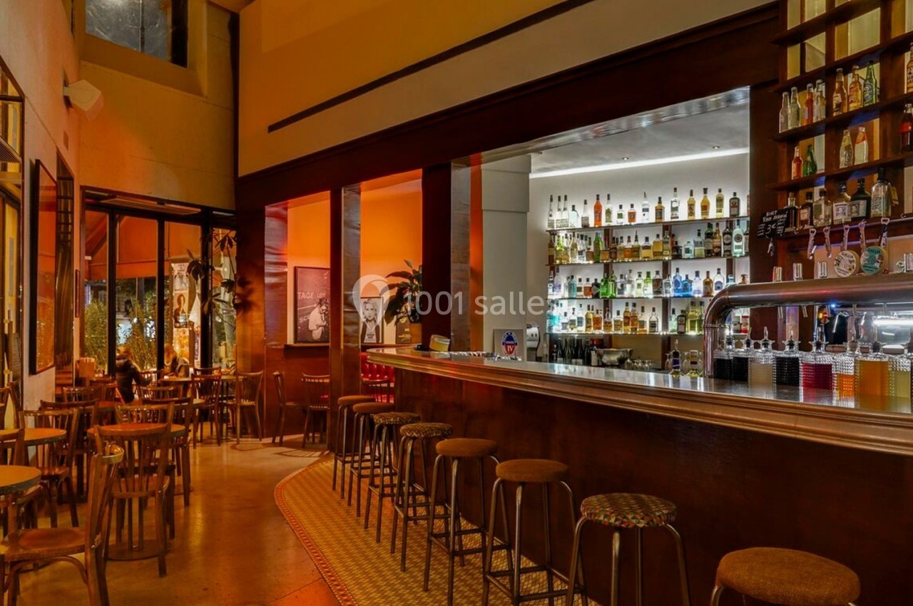 Intérieur d'un bar avec un long comptoir, des tabourets alignés et des étagères remplies de bouteilles.