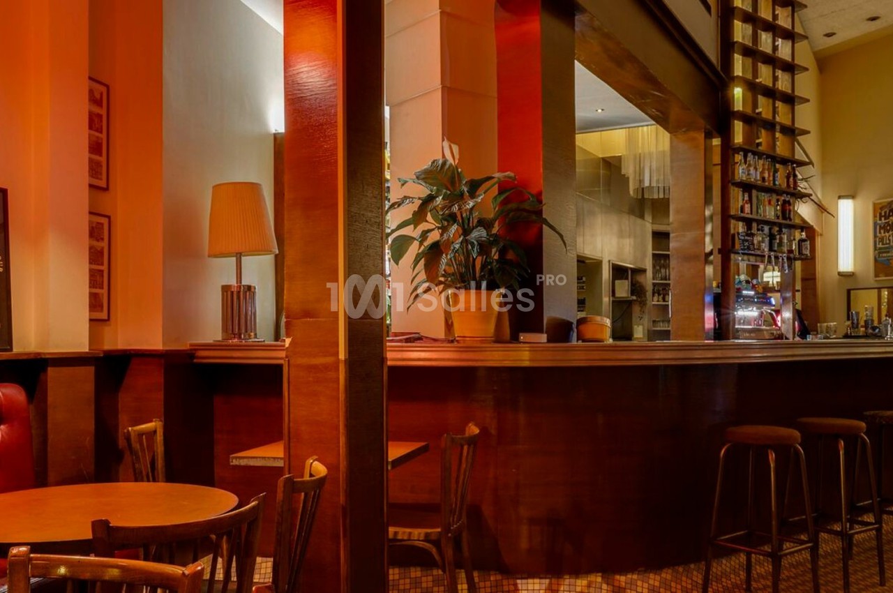 Intérieur d'un café chaleureux avec comptoir en bois, tabourets hauts, tables rondes et éclairage tamisé.