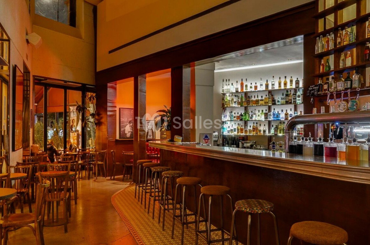 Intérieur d'un bar avec un long comptoir, des tabourets alignés et des étagères remplies de bouteilles.