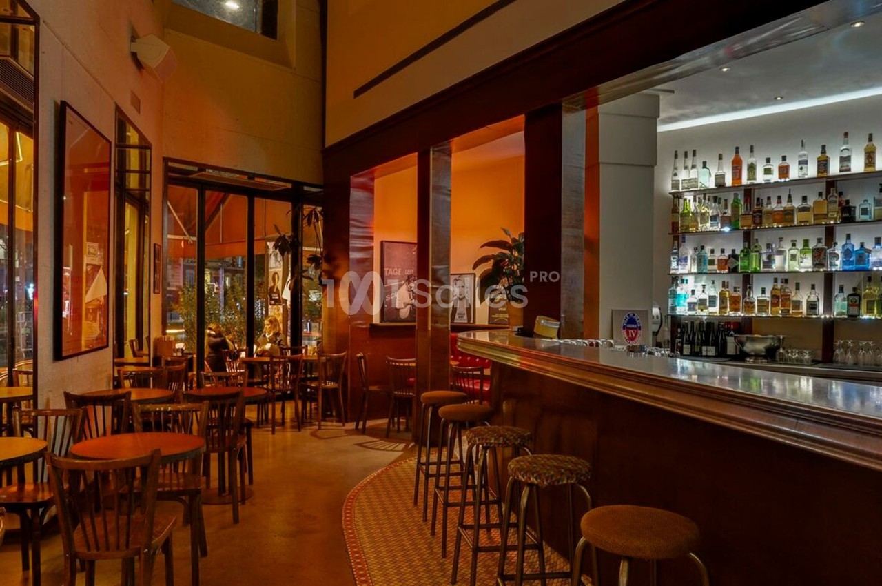 Intérieur d'un bar chaleureux avec des tables en bois, un comptoir éclairé et une étagère garnie de bouteilles.