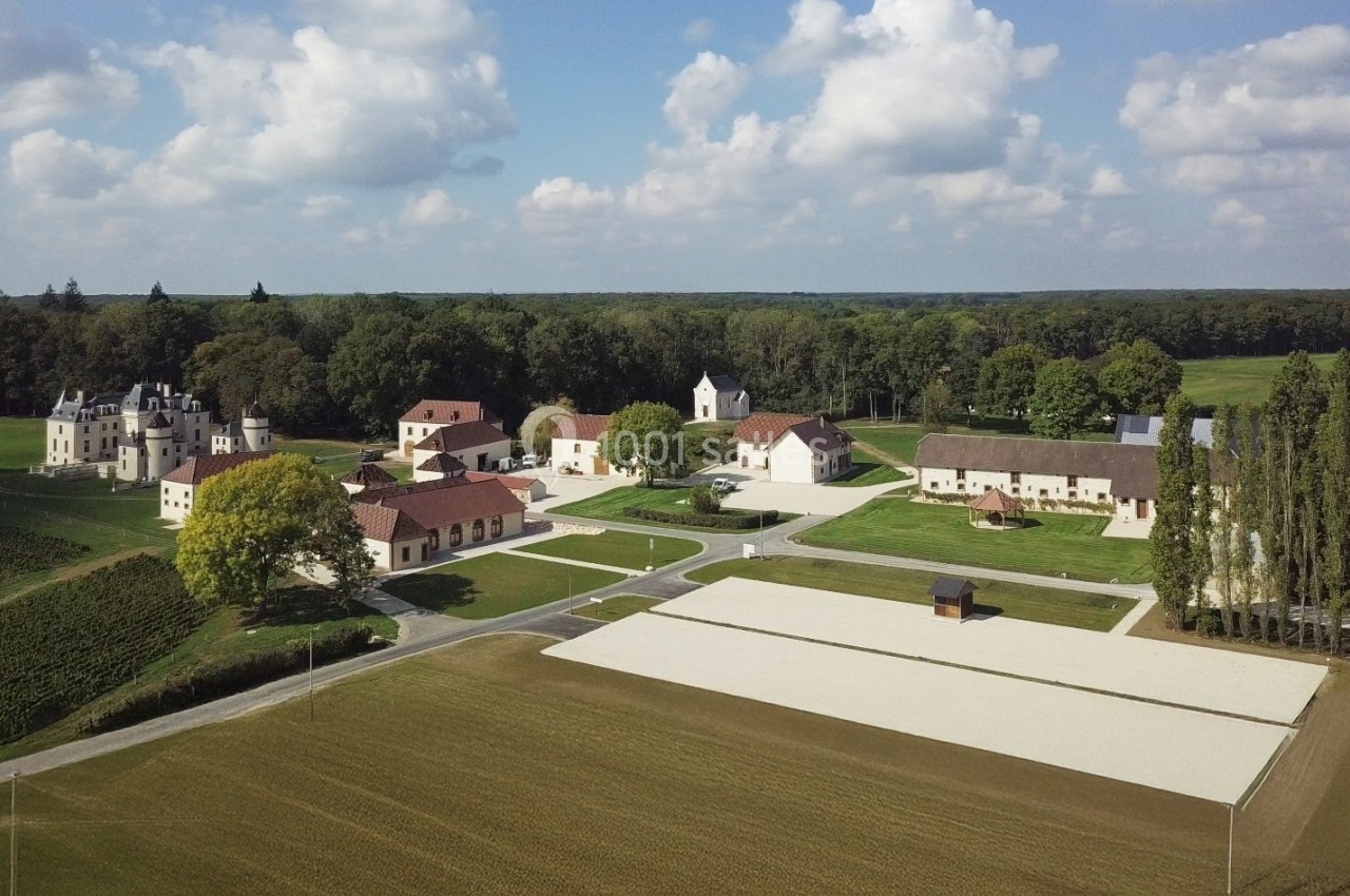 Vue aérienne d'un domaine rural avec bâtiments anciens, champs cultivés et forêt en arrière-plan sous un ciel partiellement…