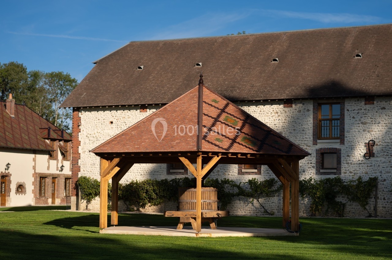 Abri en bois avec toit en tuiles dans une cour verdoyante, devant un bâtiment en pierre à colombages.