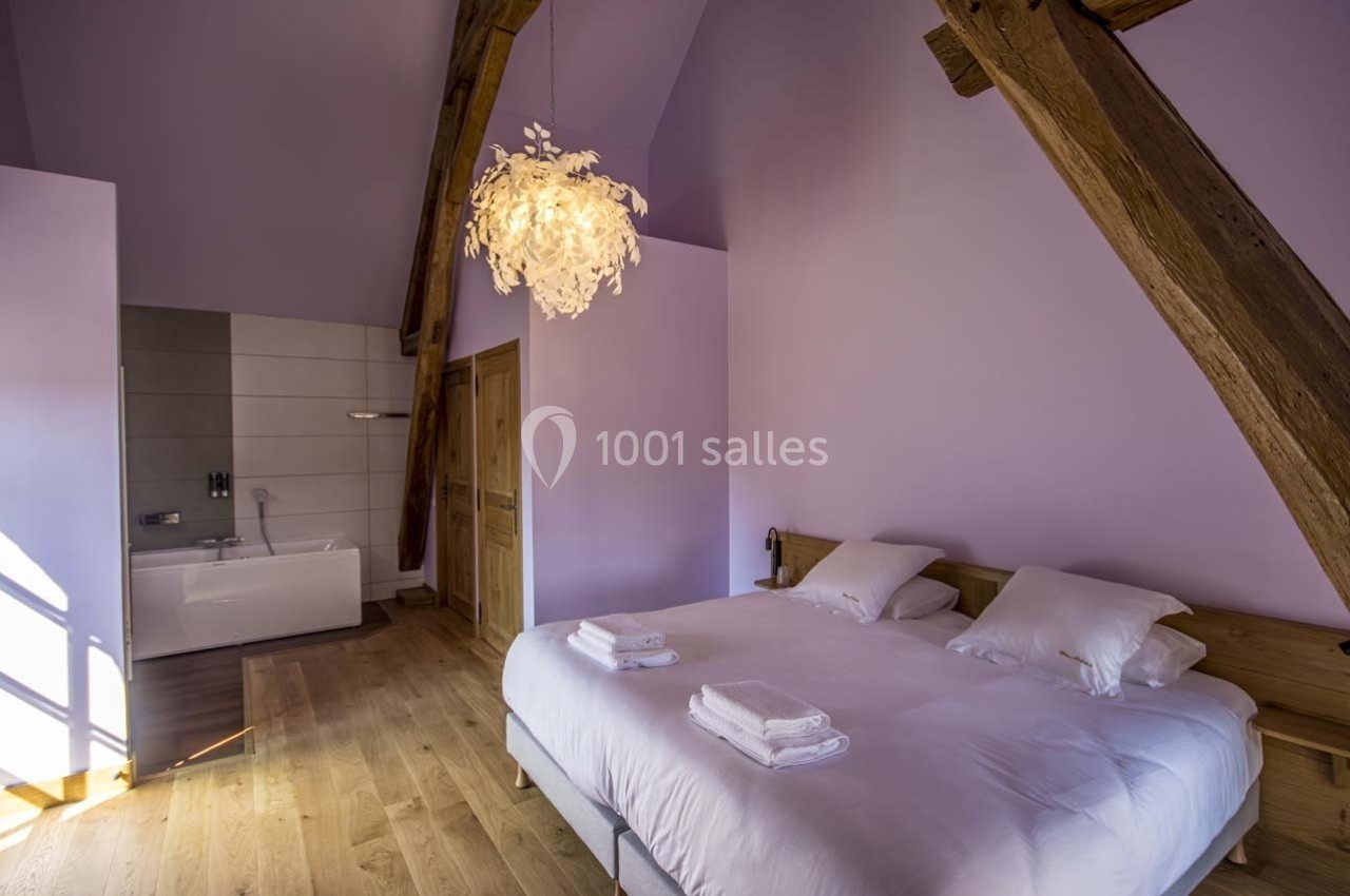 Chambre avec lit double, poutres apparentes, parquet en bois, mur lavande et salle de bain attenante avec baignoire.