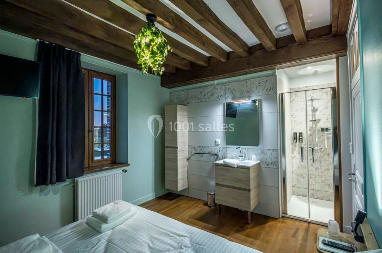 Chambre avec lit double, plafond à poutres apparentes, salle de bain attenante moderne et fenêtre avec rideaux sombres.