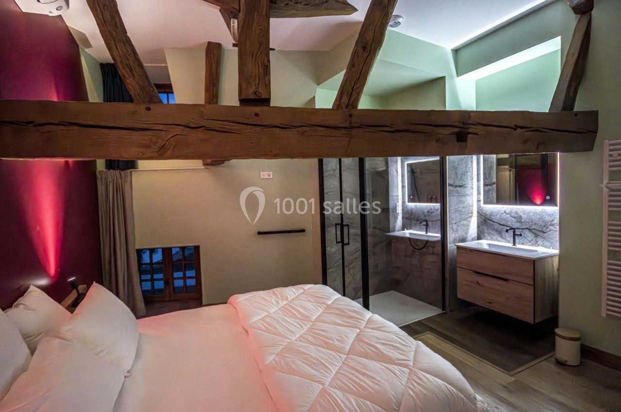 Chambre avec poutres apparentes, lit double, éclairage tamisé et salle de bain moderne attenante avec douche.
