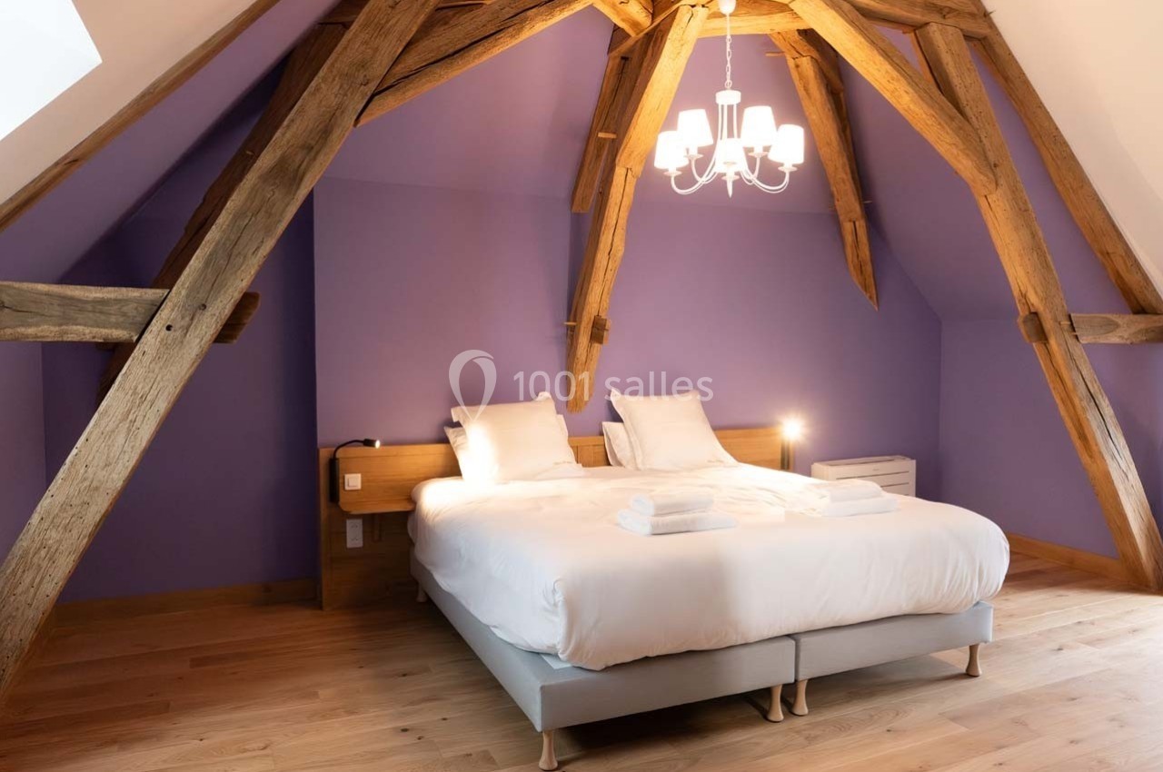 Chambre mansardée avec poutres apparentes, lit double, linge de lit blanc et mur violet, éclairée par un lustre.