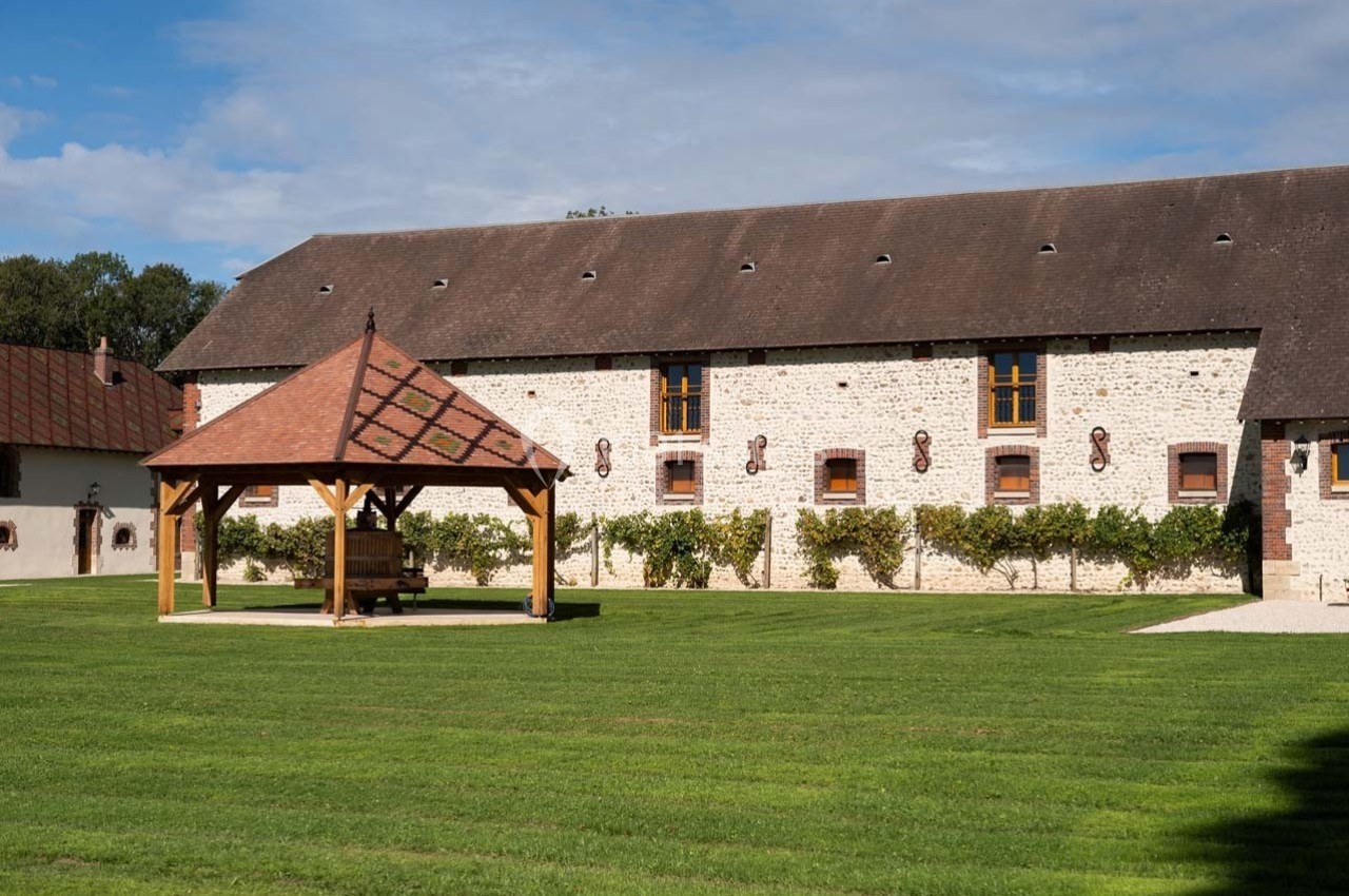 Bâtiment en pierre avec toit en ardoise, pergola en bois et pelouse bien entretenue sous un ciel dégagé.