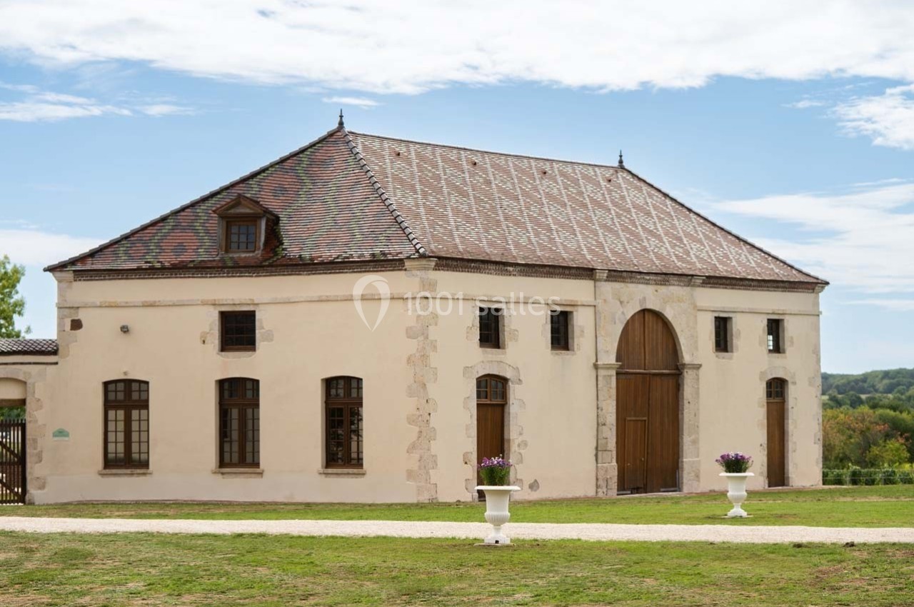 Bâtiment ancien avec toit en tuiles, grandes portes en bois et fenêtres, entouré de pelouse et de pots de fleurs.