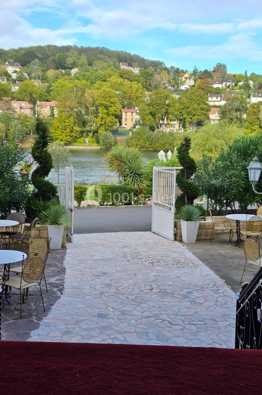Vue sur une terrasse avec des tables et des chaises, donnant sur un jardin, une rivière et des collines verdoyantes.