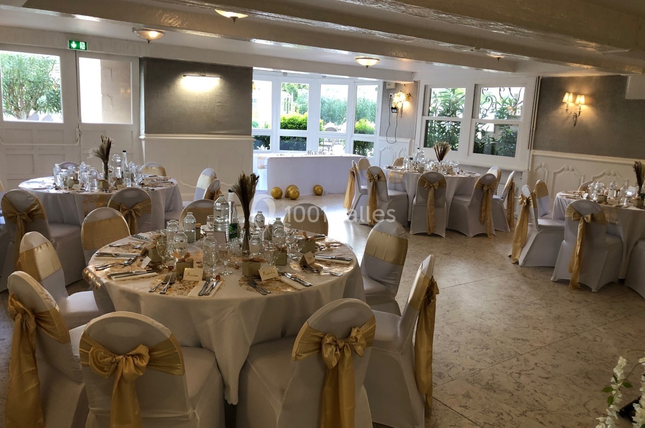 Salle de réception décorée avec des tables rondes dressées, nappes blanches et chaises ornées de nœuds dorés.