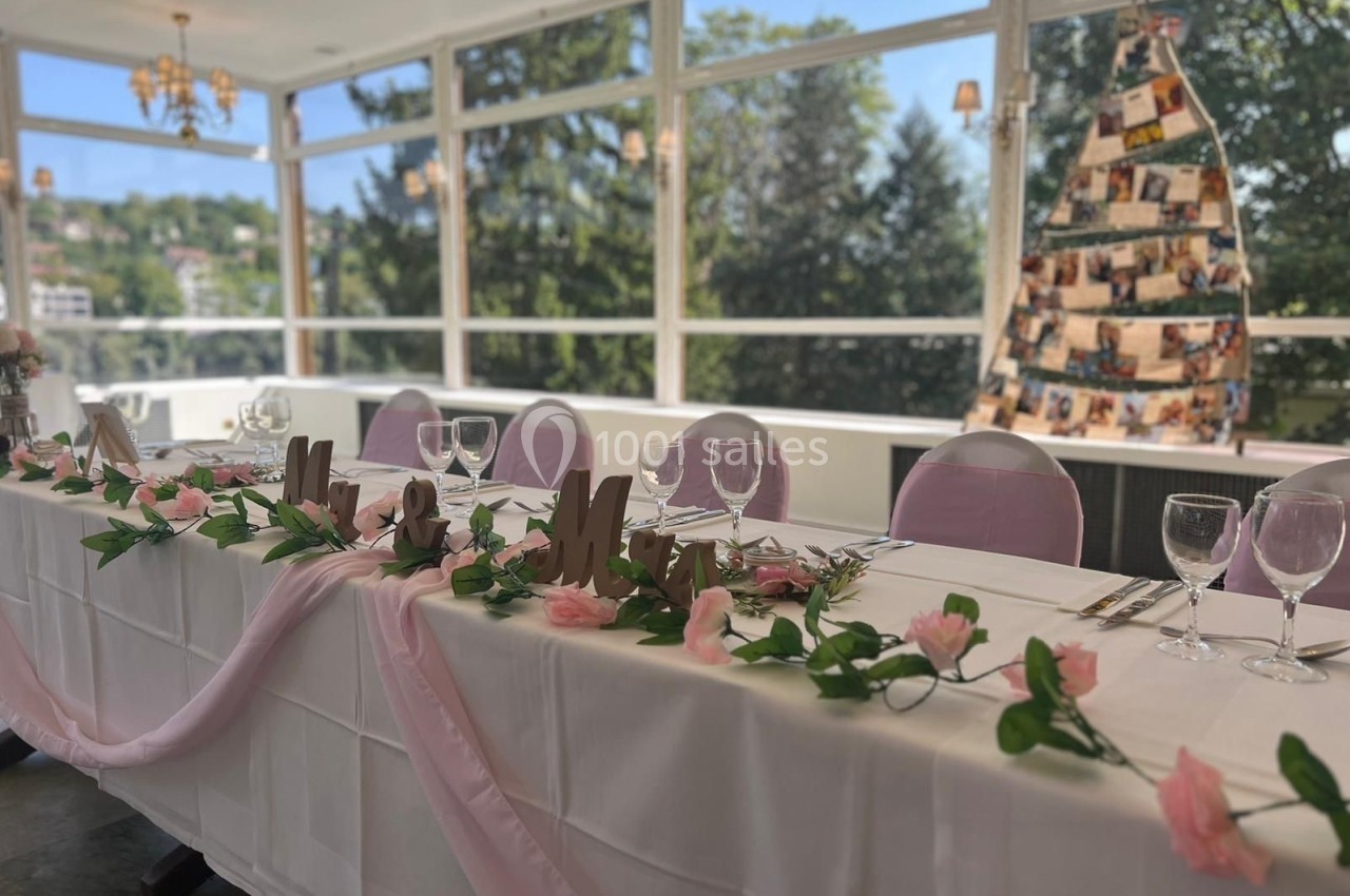 Table décorée pour un événement avec nappes blanches, fleurs roses, verres et vue sur un paysage arboré en arrière-plan.