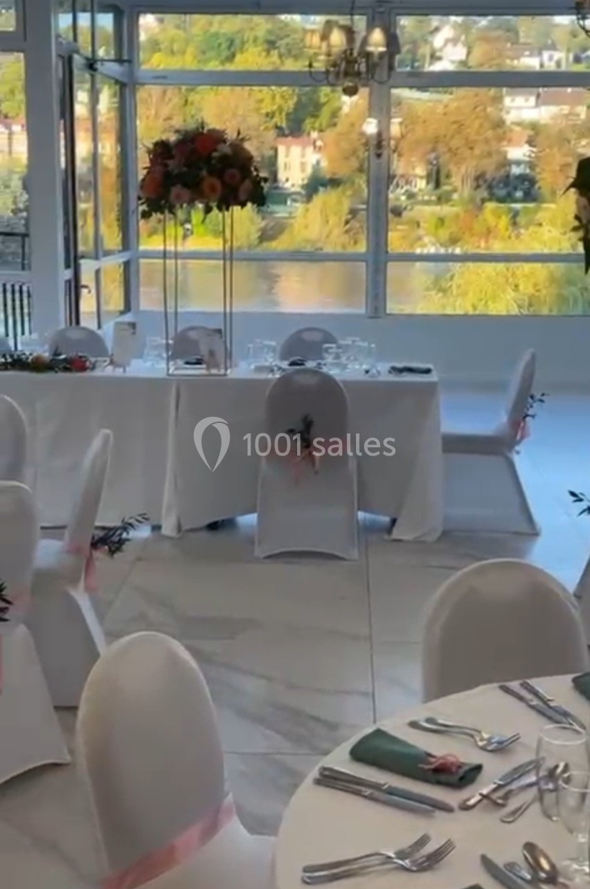 Salle de réception lumineuse avec tables dressées, chaises décorées et vue sur un paysage naturel à travers de grandes baies…