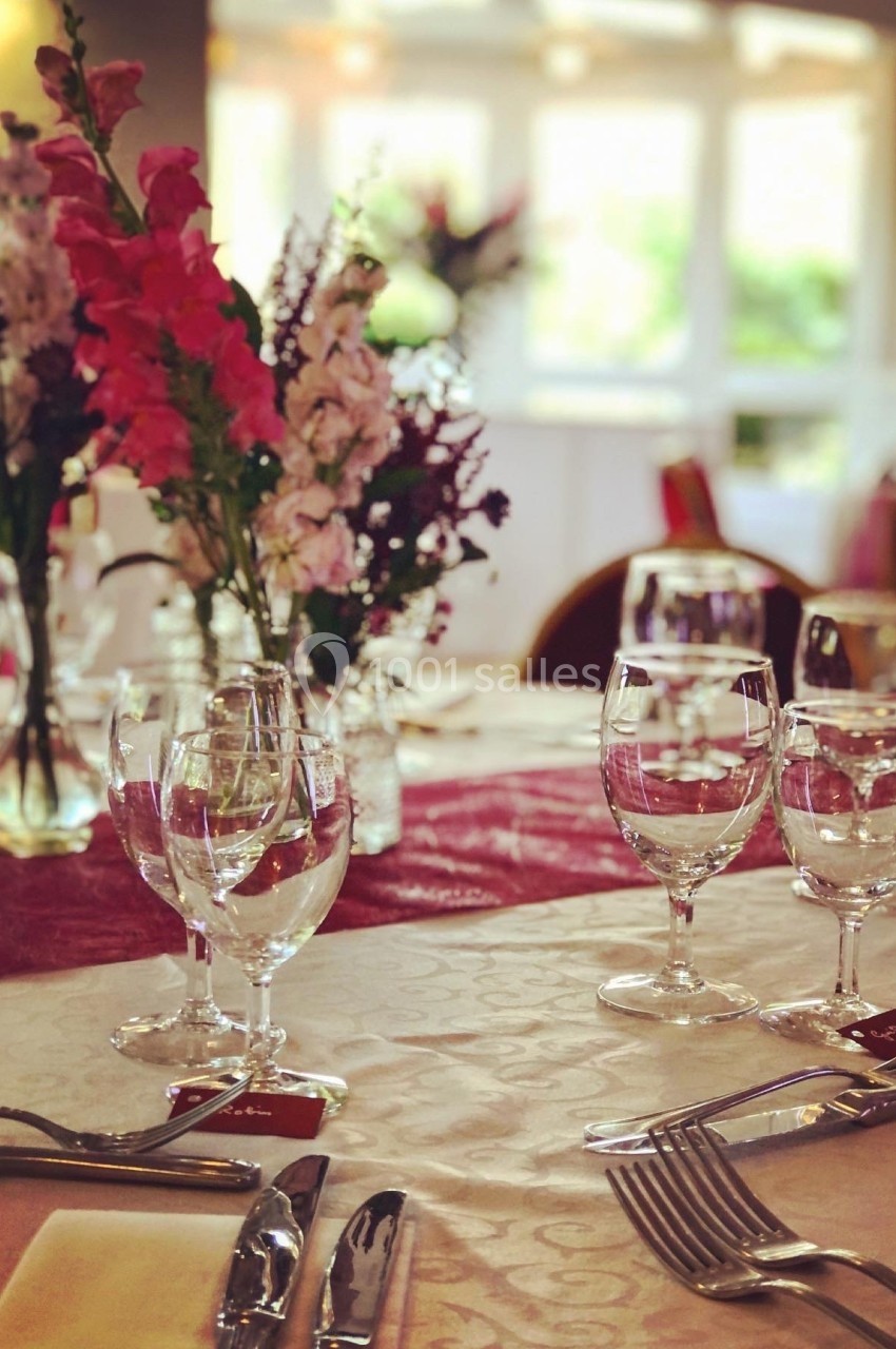 Table élégamment dressée avec nappes blanches, verres, couverts et bouquets de fleurs colorées en arrière-plan.